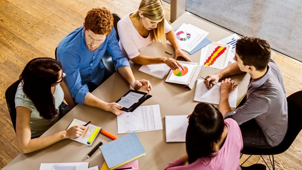 上から見た図。机を囲んで座り、書類やタブレットを見ながら話し合っている5人のグループ。