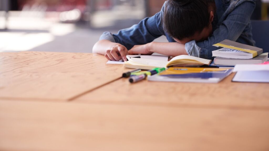 木製の机の上で、たくさんの本やマーカーに囲まれながら突っ伏して眠っている人。