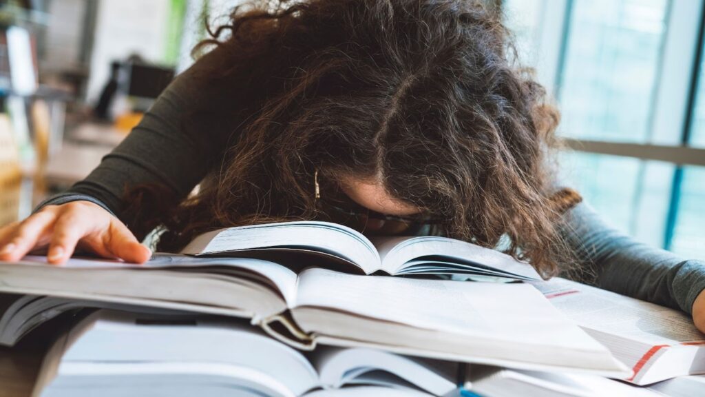 山積みの本の上に顔をうずめて、疲れ果てて眠っている女性。