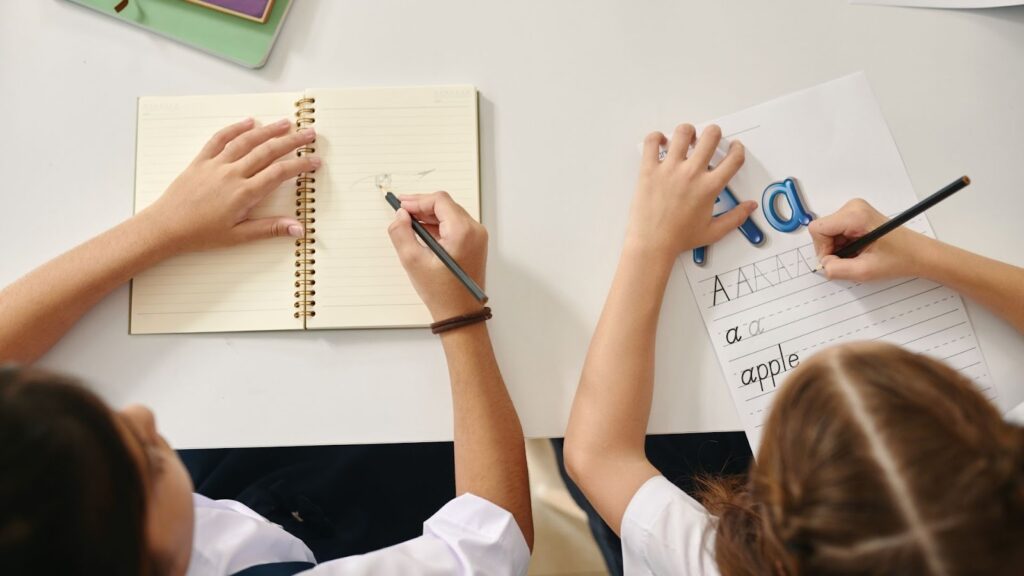 上から見た図。2人の子供が机に座り、一人はノートに絵を描き、もう一人はアルファベットの練習をしている。